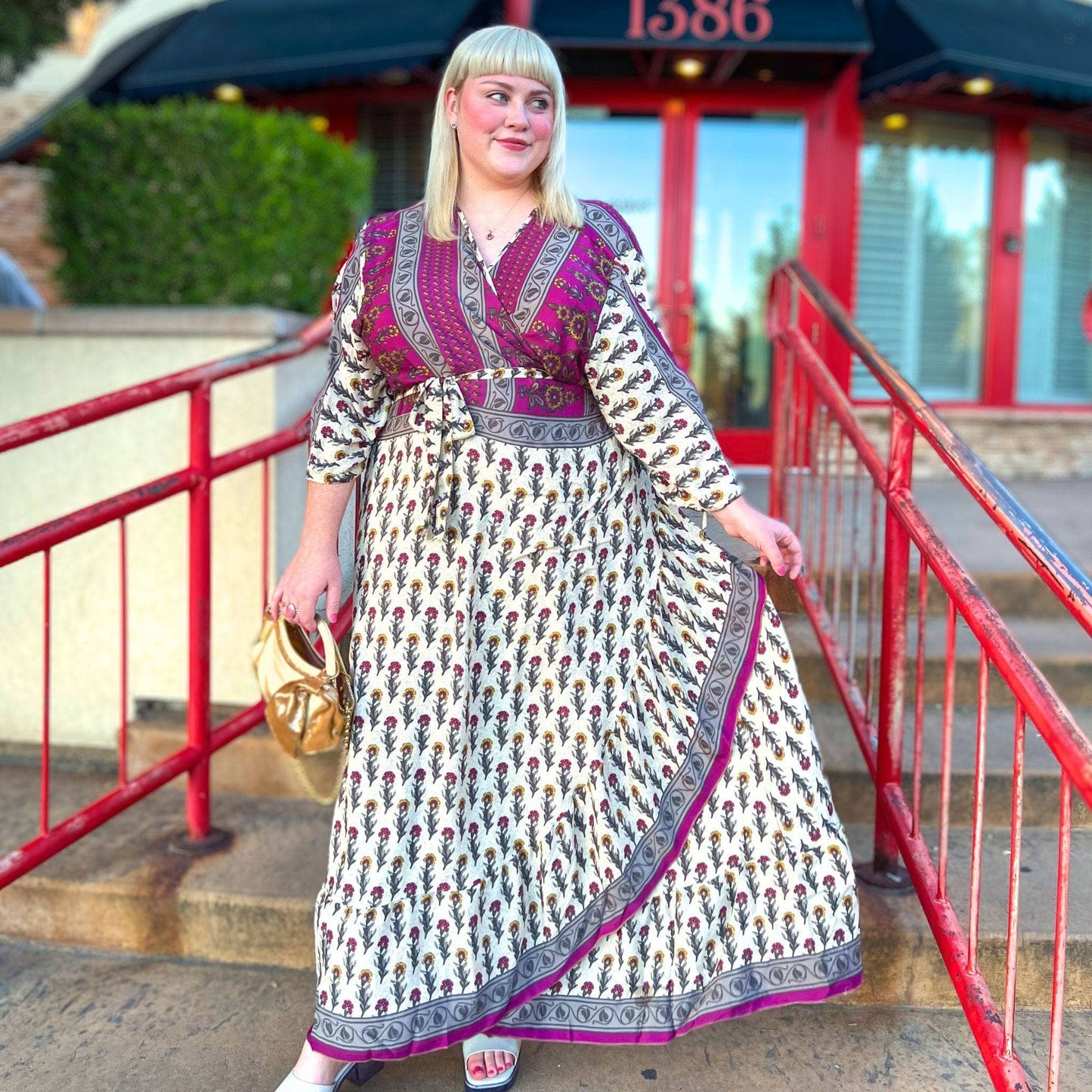 A model standing on the steps outside of a restaurant. She's wearing a reclaimed sari wrap dress paired with a while leather shoe. The wrap dress has a magenta top with a white bottom and grey edges. There's a magenta floral pattern across the whole dress.