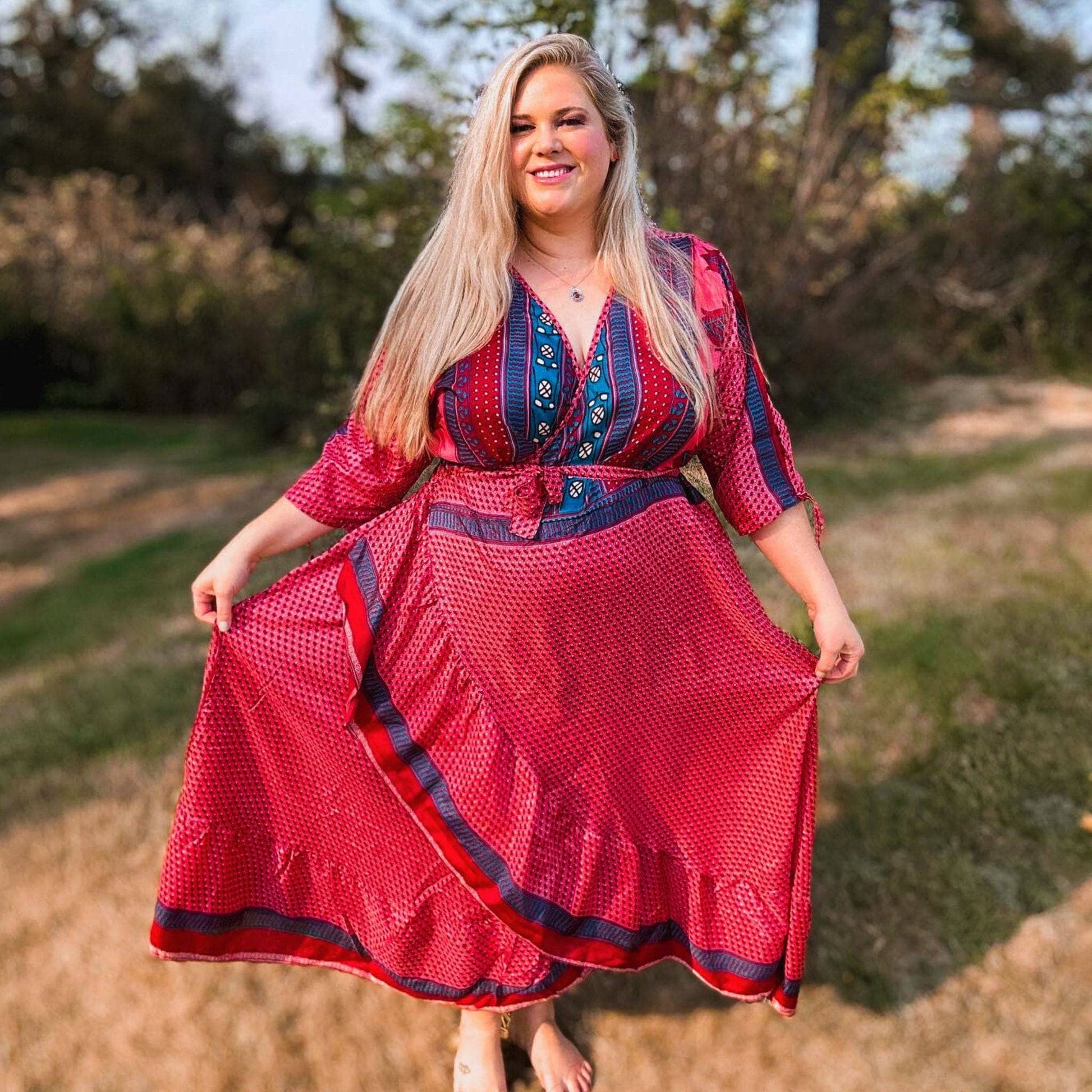 A woman standing in a field wearing a a red sari silk wrap dress with navy polka dots across it. The top of the dress has light blue stripes with floral embellishments.