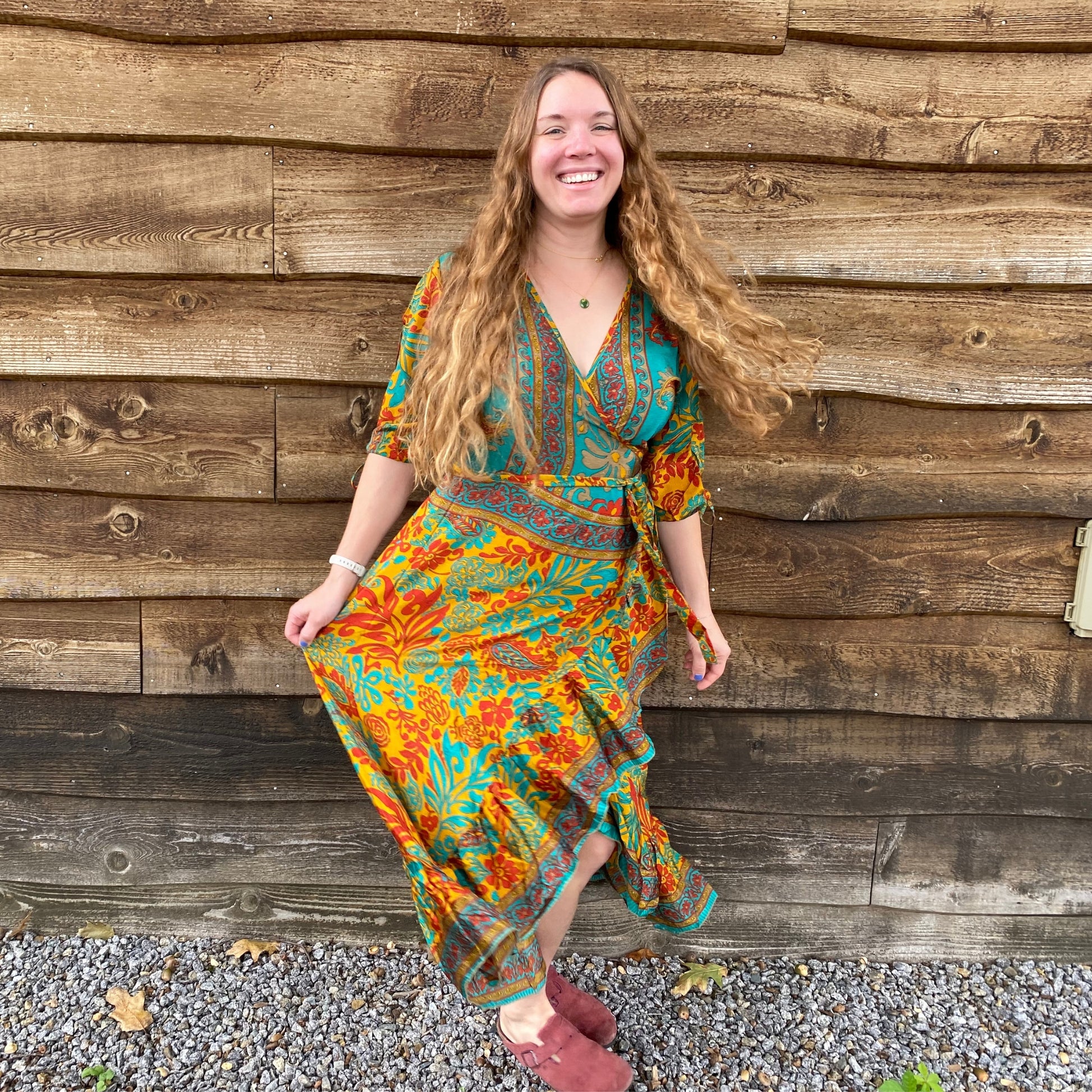 A Model standing against a wooden house wearing a warmer color Zaria Wrap Dress. The Dress is Mustard Yellow, Red and Light Green with floral patterns..