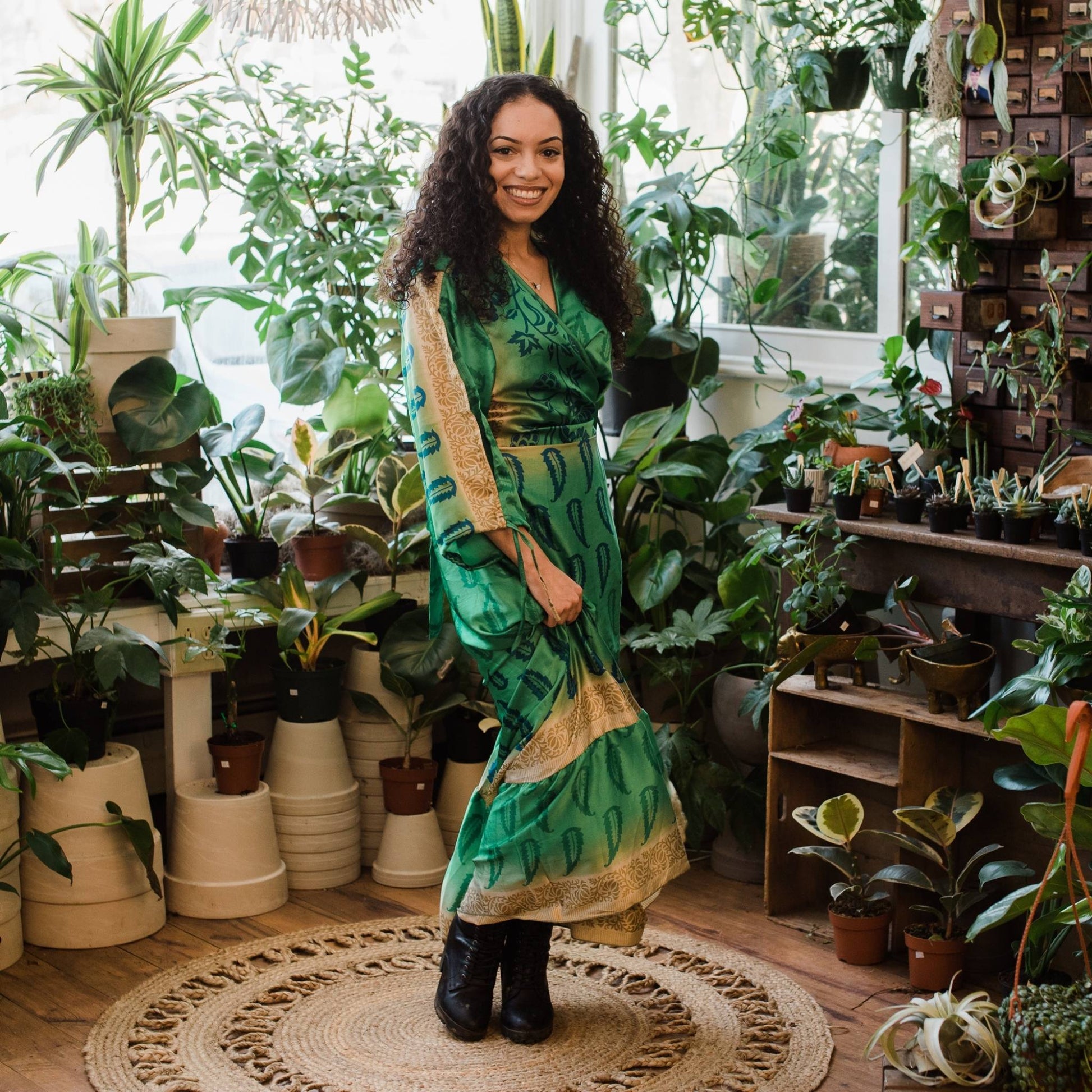 Model is standing a room with assorted plants wearing a green Zaria wrap dress in our women's size.