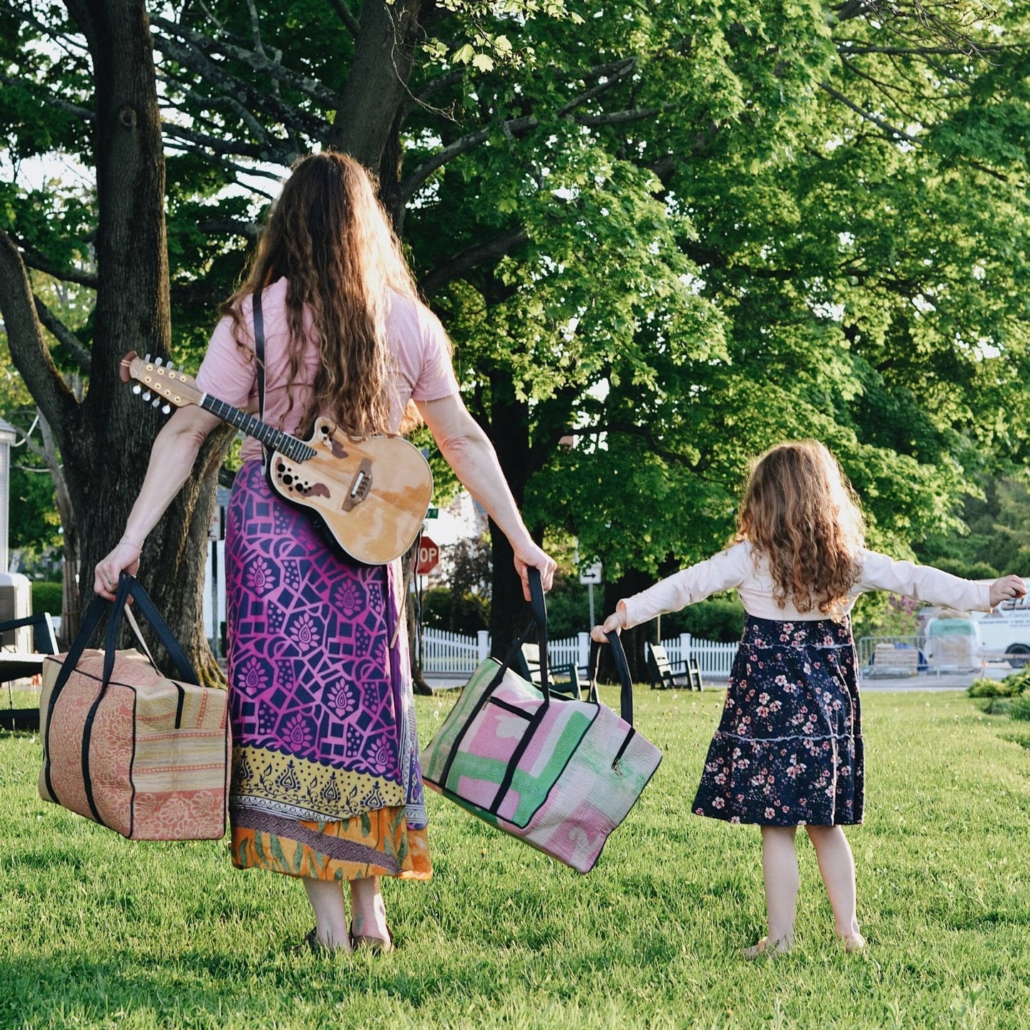 the backs of an adult model and a kid model, both holding a kantha duffle bag with their arms stretched out. while standing in the park.