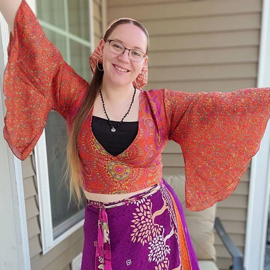 Model wearing orange Sari wrap top with bell sleeves paired with sari wrap skirt with exterior wall in the background.