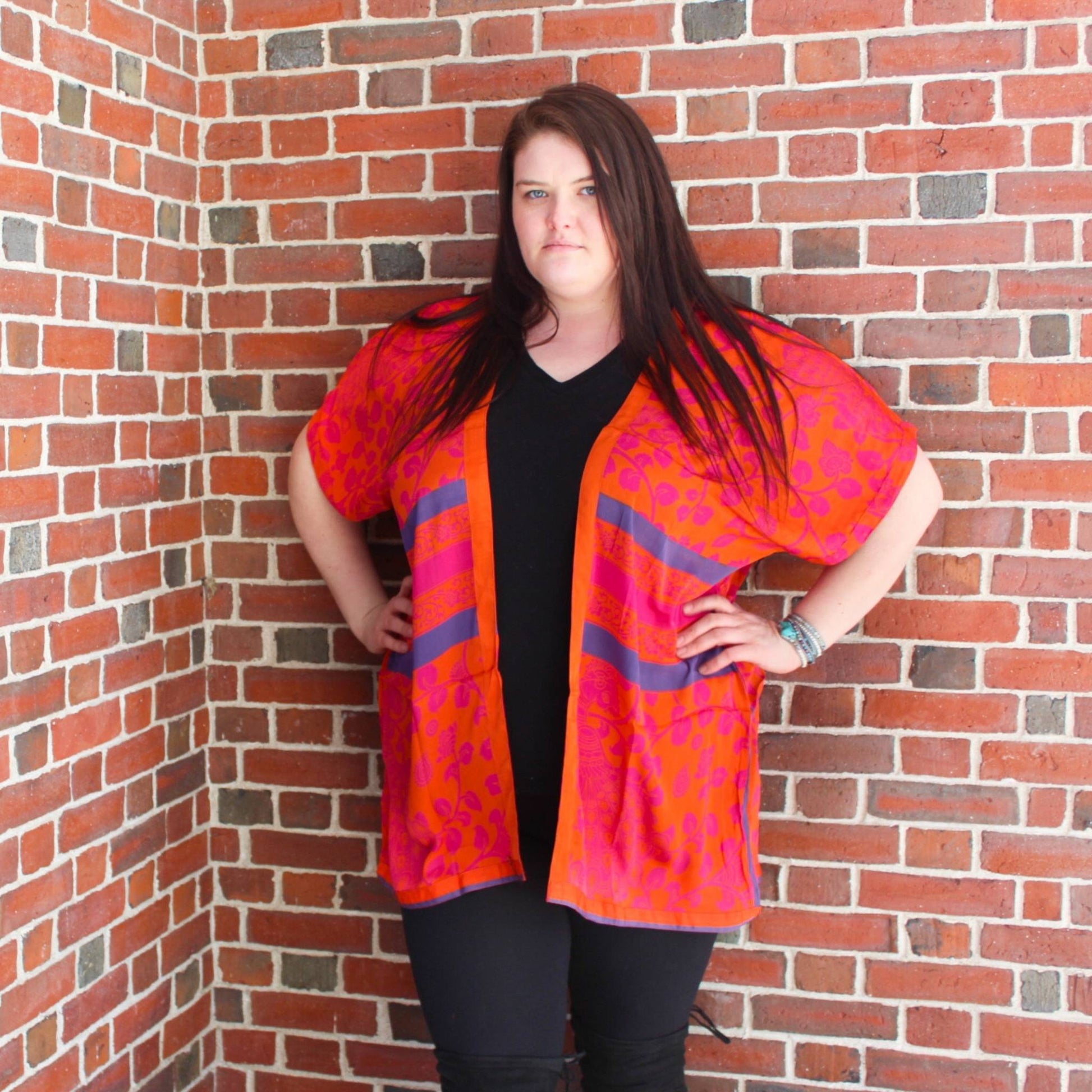 Model is standing in front of a brick wall wearing a bright orange + pink amra shirt duster in our goddess size.