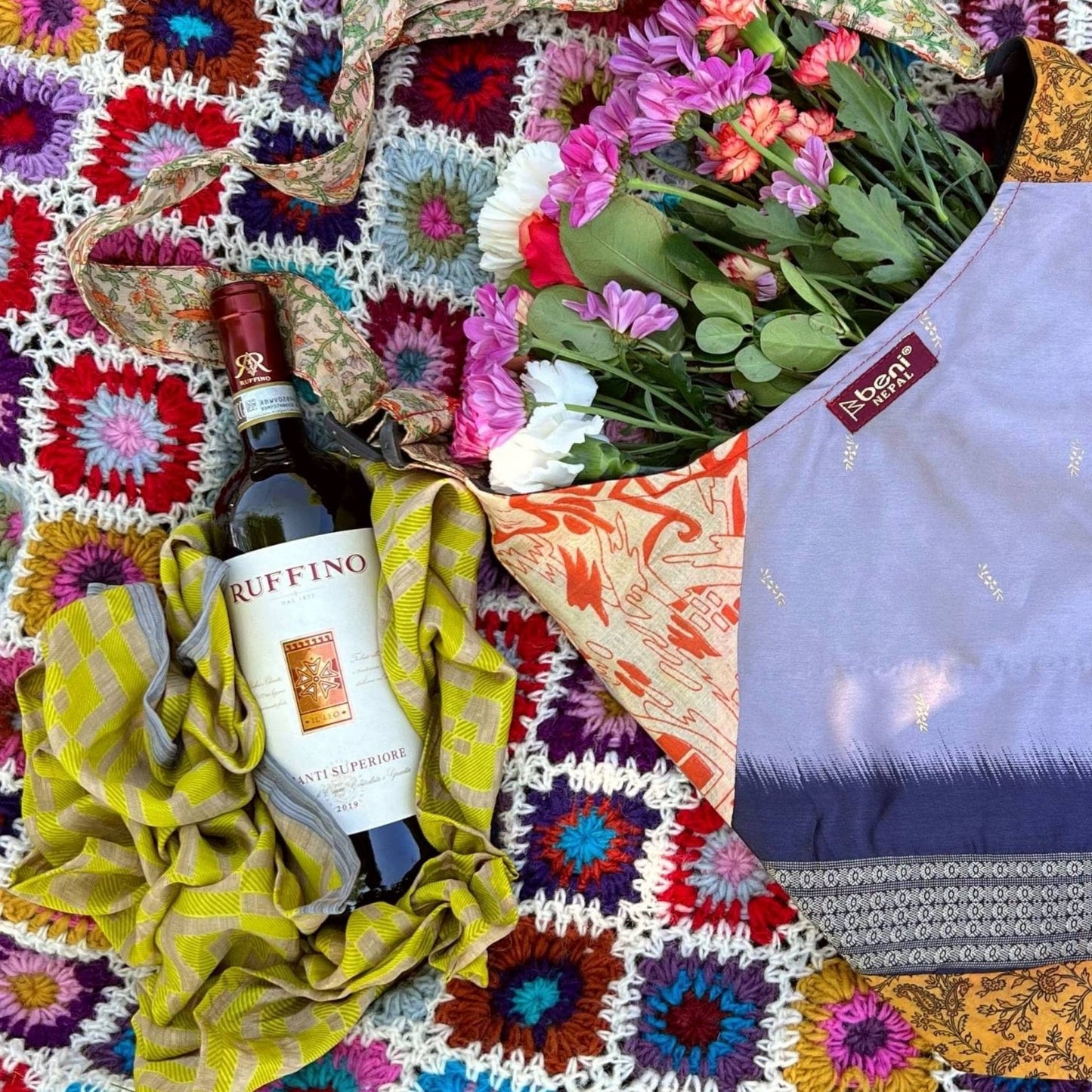 Green furoshiki is laying under a wine bottle on a crochet blanket in the park next to a sari silk purse with flowers in it.