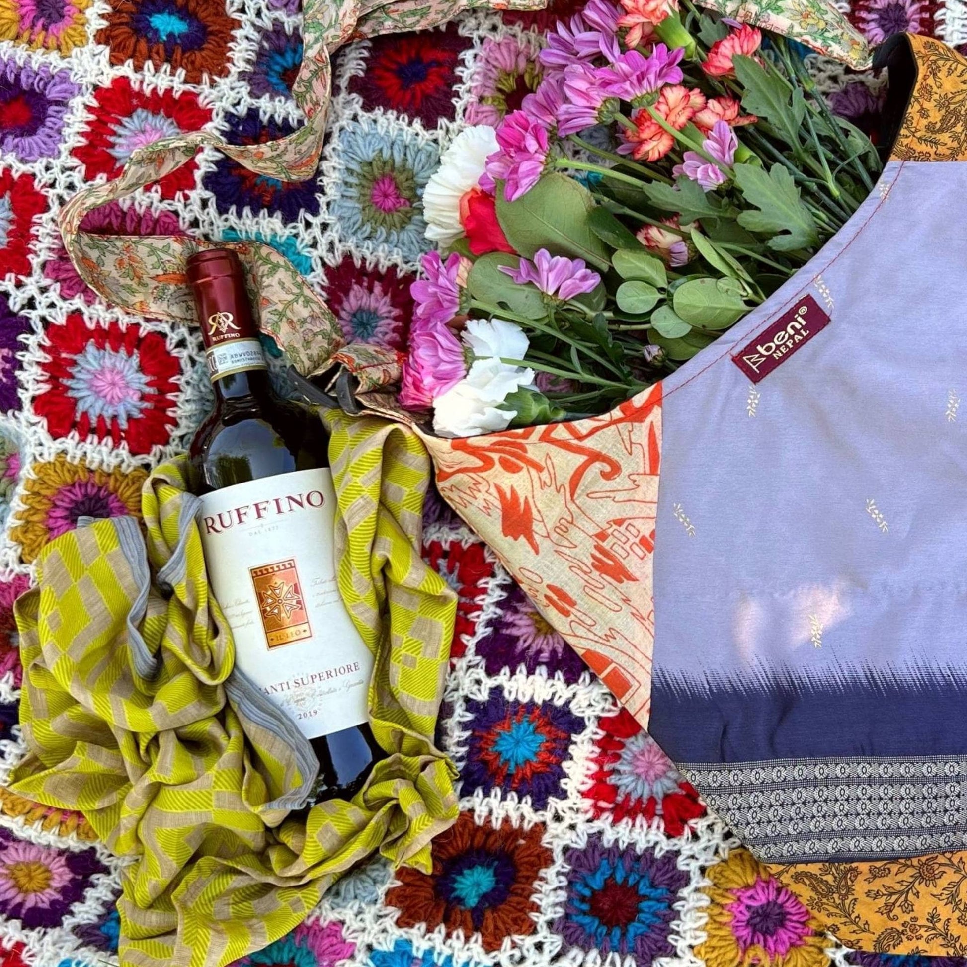 Green furoshiki is laying under a wine bottle on a crochet blanket in the park next to a sari silk purse with flowers in it.
