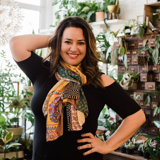 Model has one hand on their head and one on their waist wearing a yellow medley scarf while potted plants in the background. 