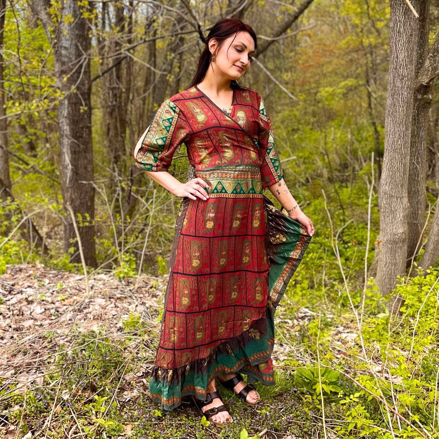 A model standing in a forest wearing a maroon sari silk wrap dress and a gladiator sandal. The dress has emerald edges and shimmering gold accents across.