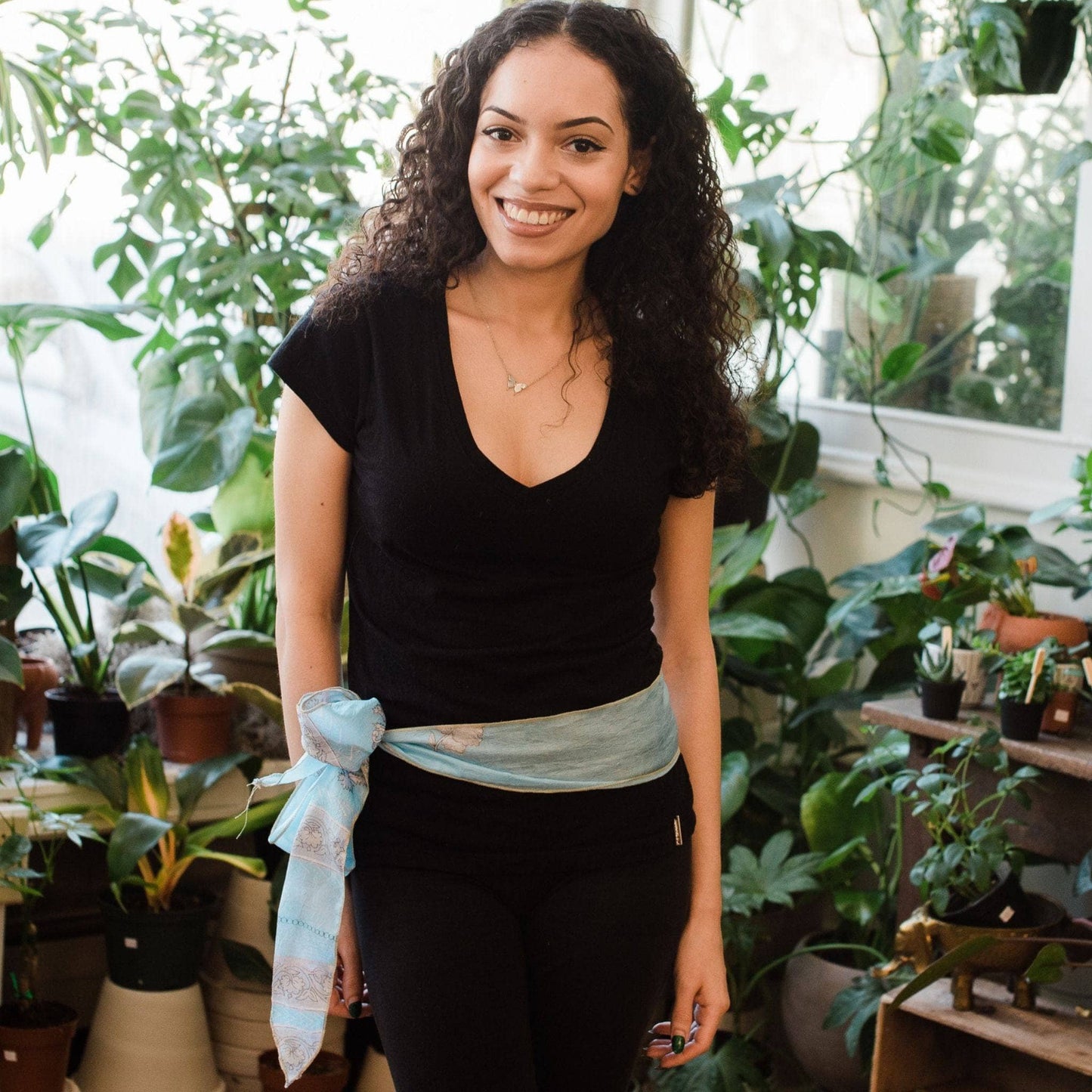 Model wearing light blue boho vintage sash accessory as a belt with a bow tied on one side with greenery in the background.