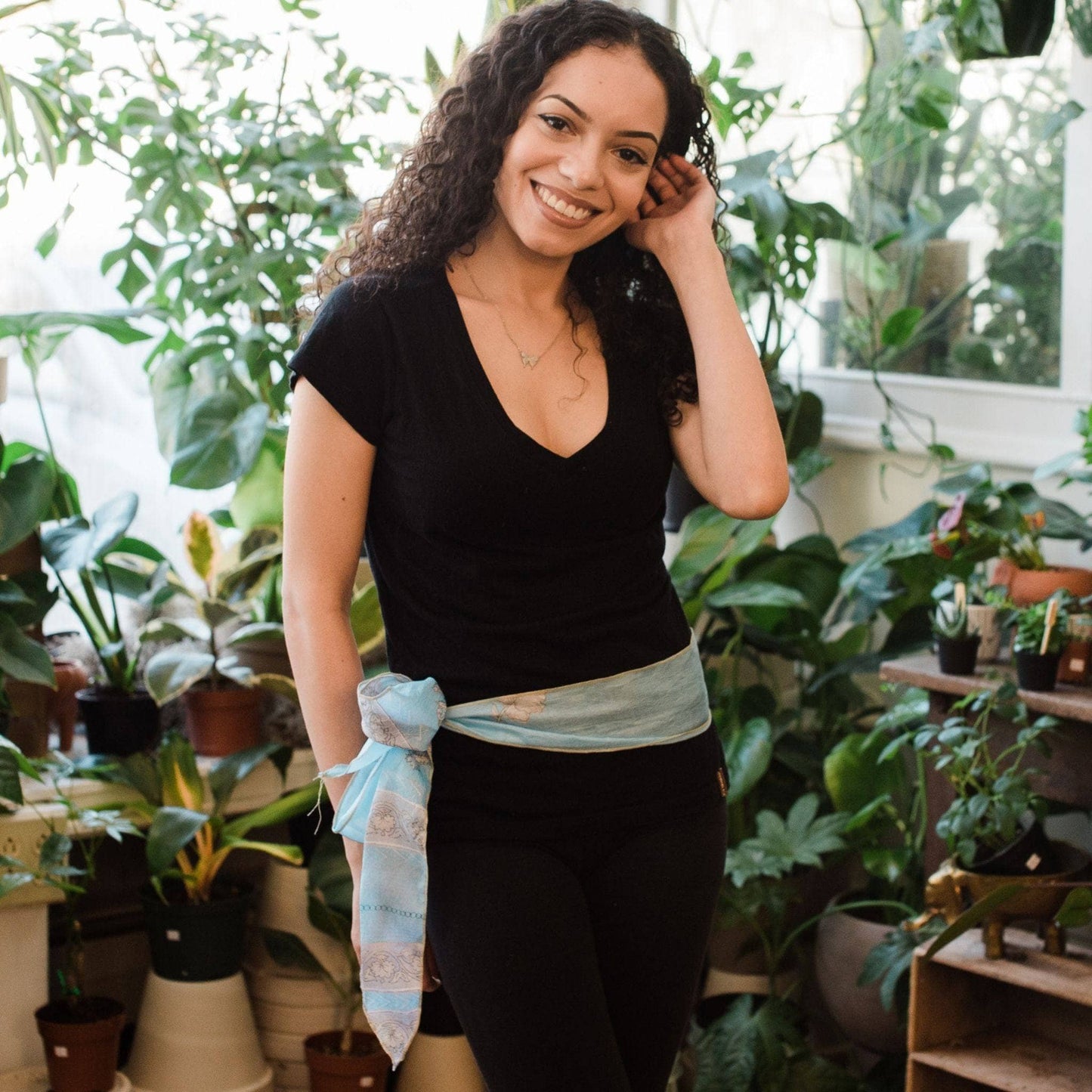 Model wearing light blue boho vintage sash accessory as a belt with a bow tied on one side with greenery in the background.