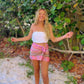Smiling blonde woman in white top and pink patterned skirt. Arms outstretched, tropical background.