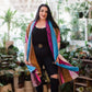 Woman in black outfit with colorful patchwork shawl. Plants in background. Smiling at camera.