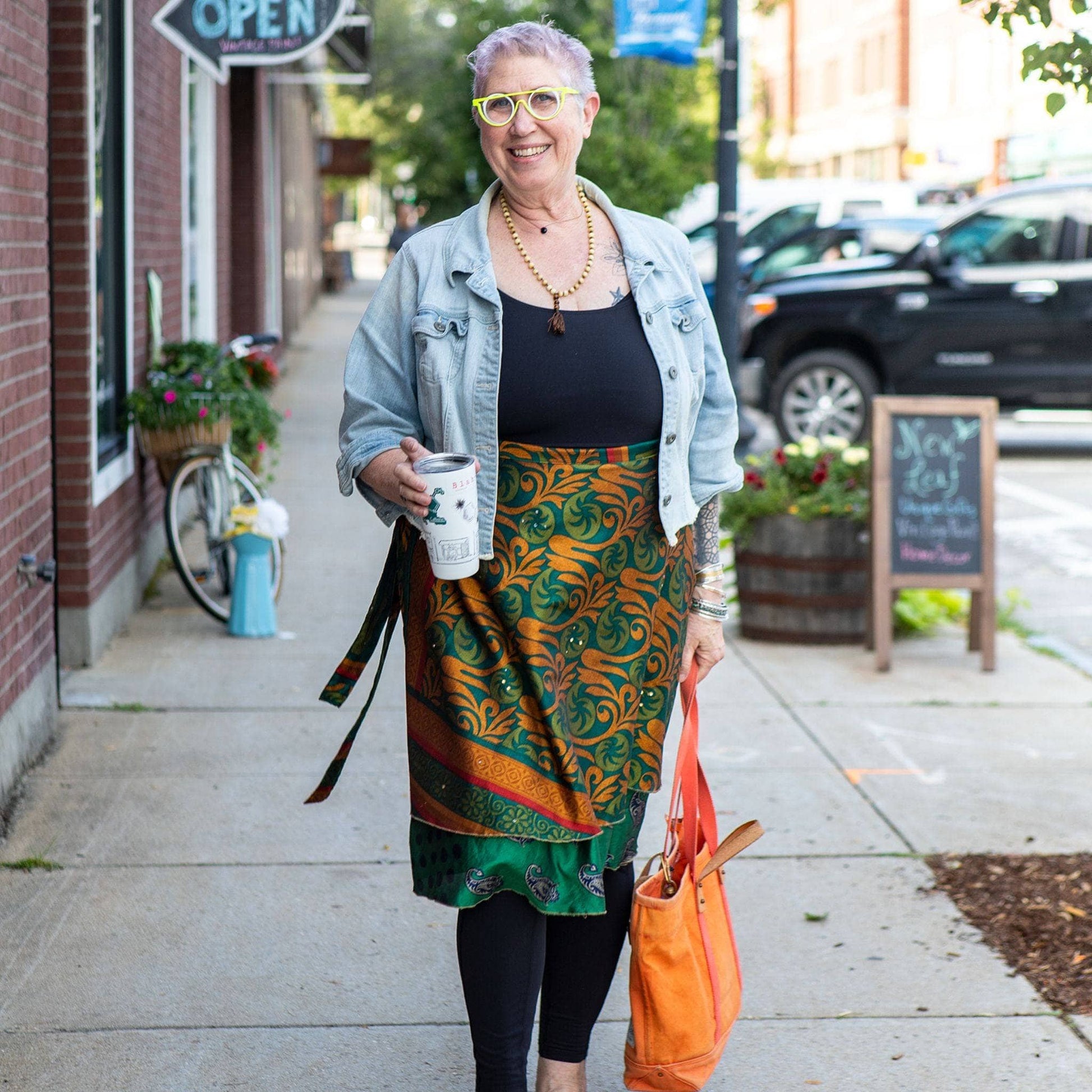 An older woman with purple hair, glasses, denim jacket, patterned skirt, holding coffee and a bag.