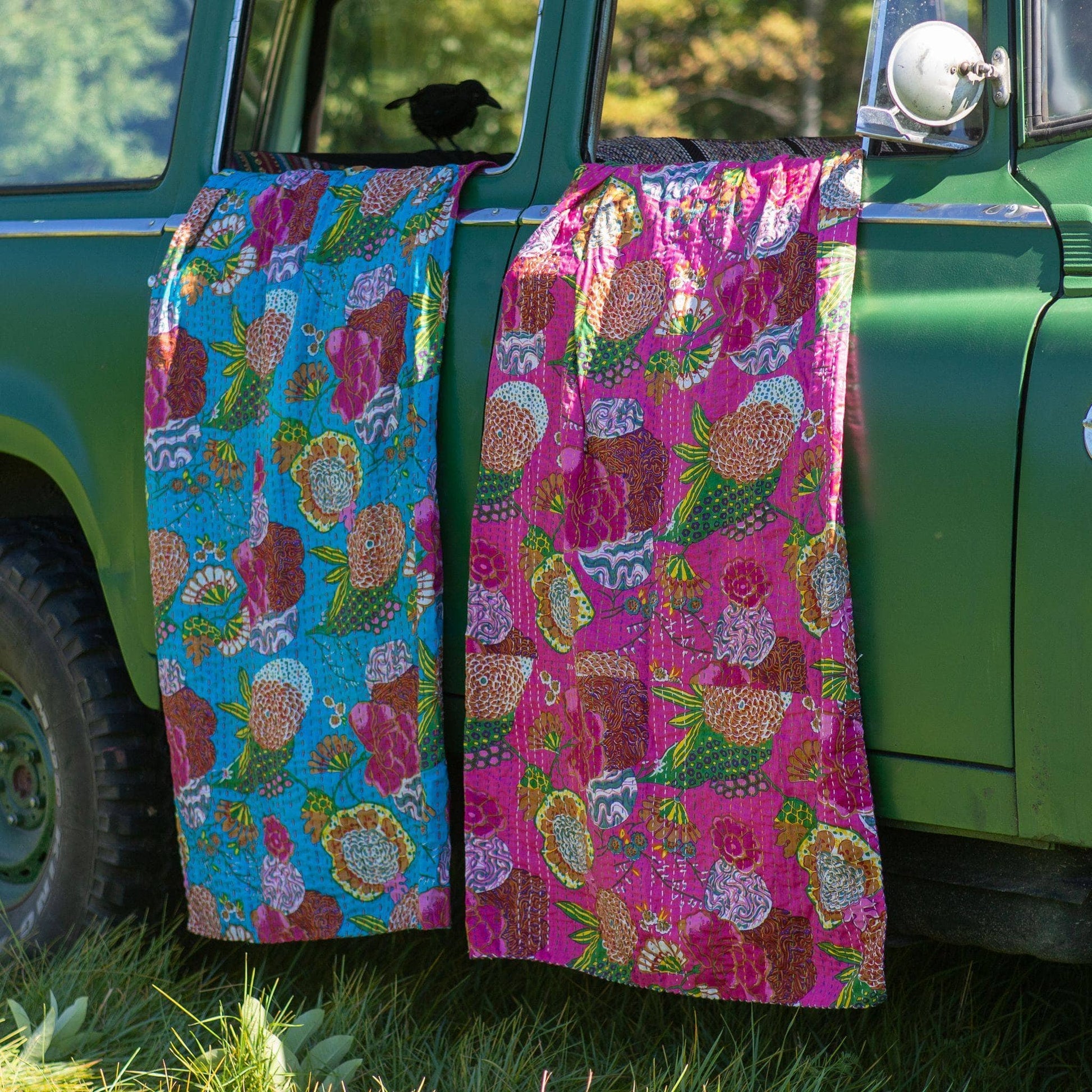 two kantha quiltshanging from a green truck's open windows. Left to right blue and pink.