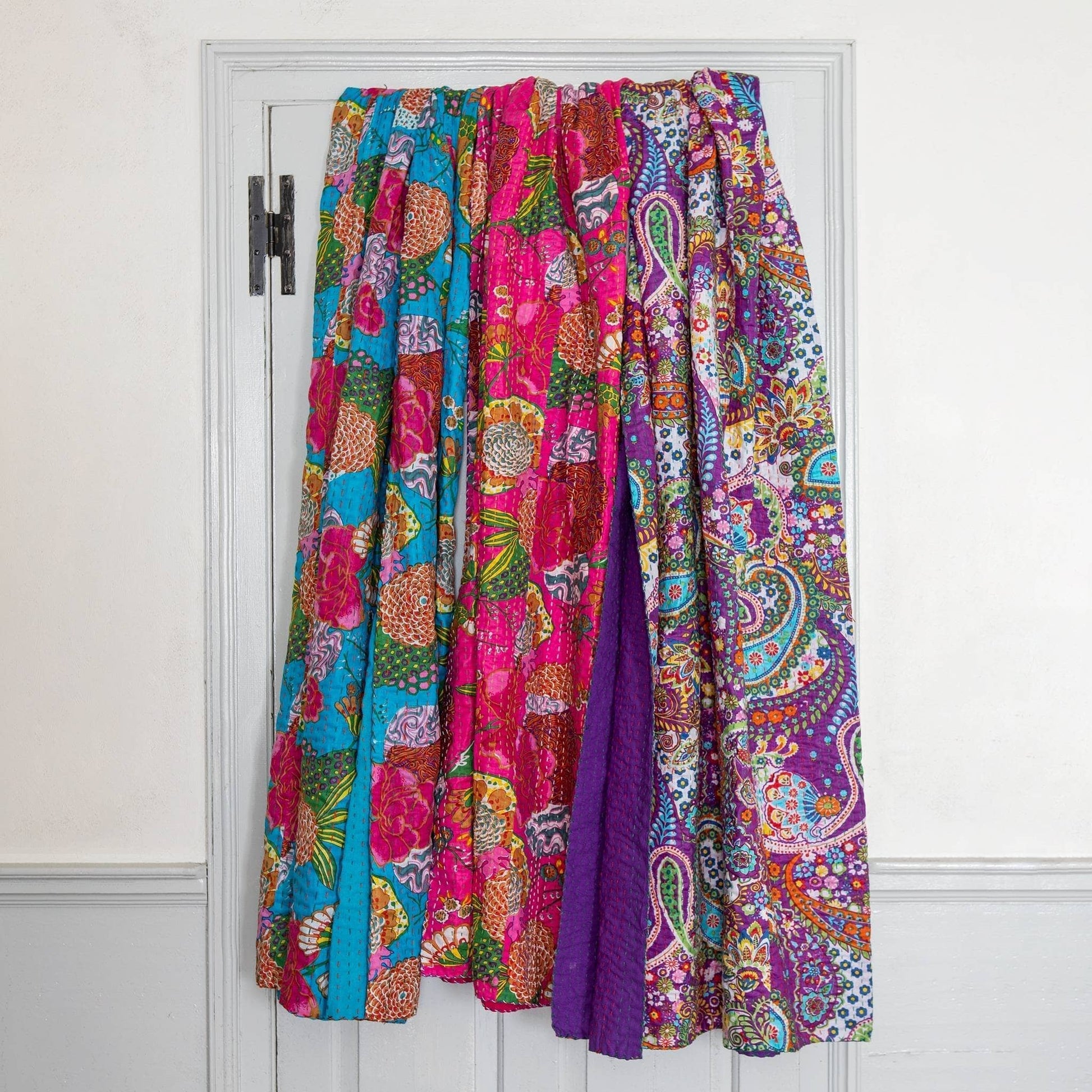 3 kantha quilts draped over the back of a white door, with a white wall surrounding. From left to right is blue, pink, and purple. 
