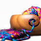 Wooden brown yarn bowl with swirl cutout for yarn ends filled with multicolored yarn and sitting on a white surface