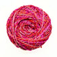 one skein of journey recycled silk yarn in the Field of Poppies colorway (variegated pink, orange, red) in front of a white background.