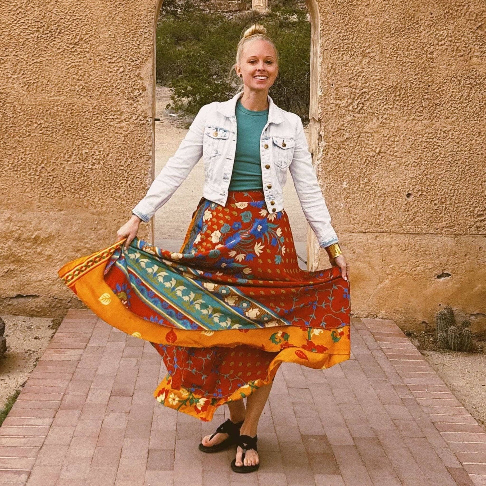 Person on a brick path, smiling in denim jacket and colorful skirt, with an arched doorway behind.