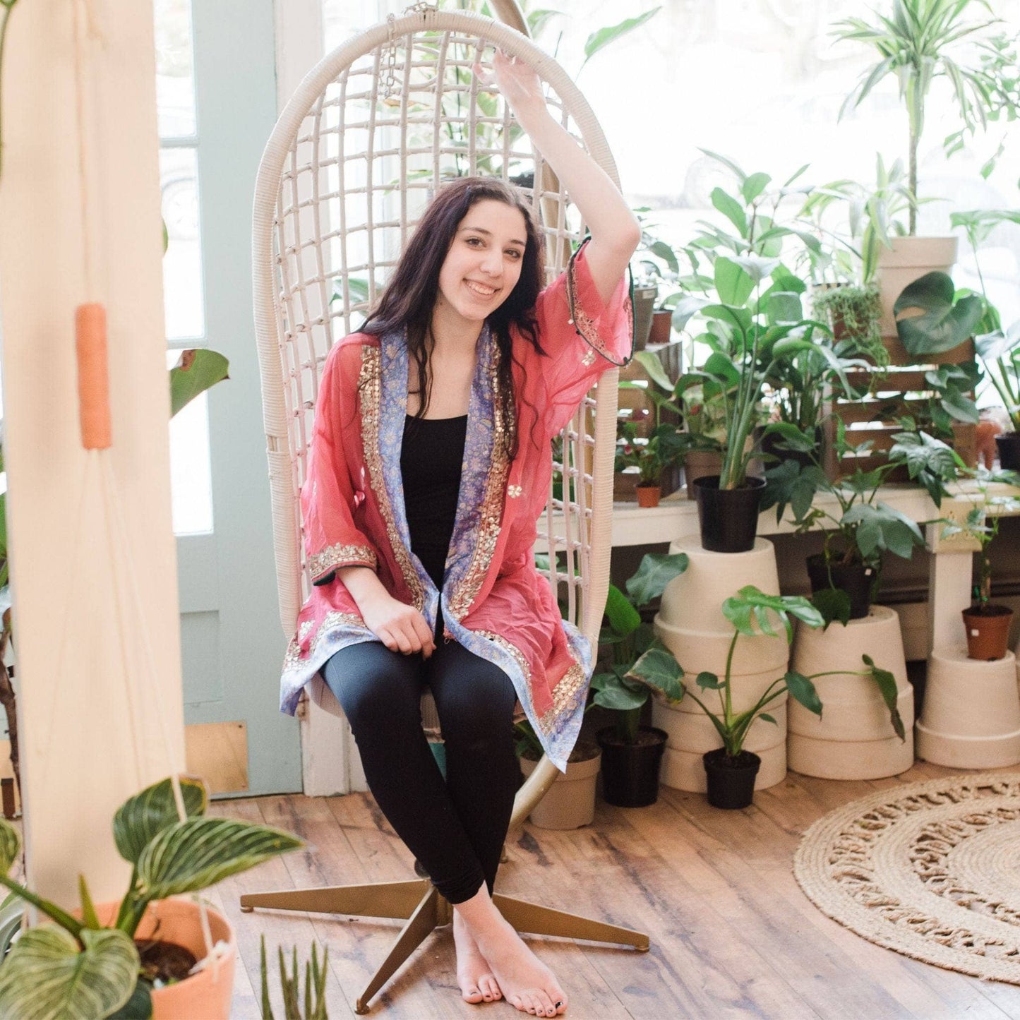 Petite model is wearing a peach and purple sophia silk duster while sitting in a chari swing with potted plants in the background.