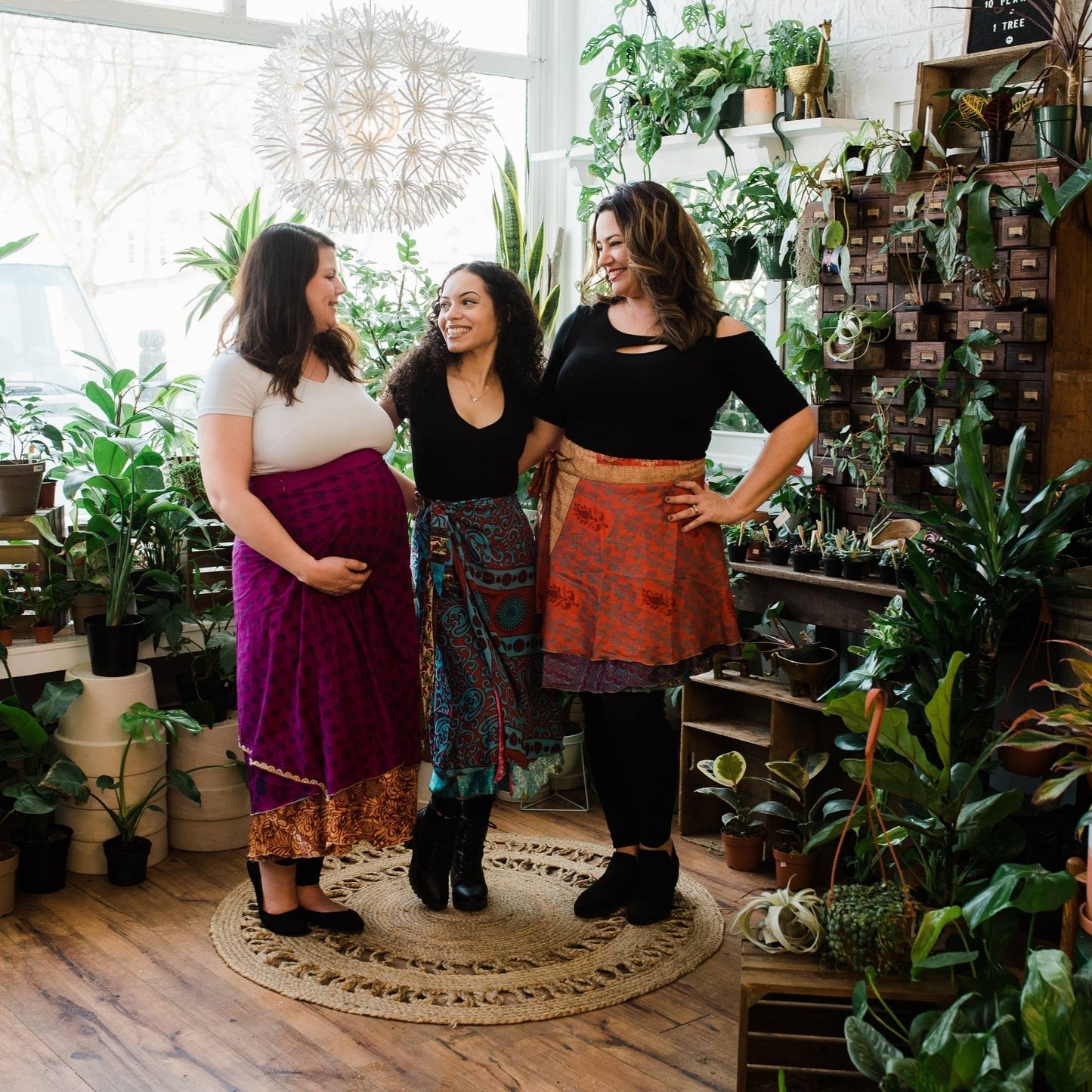 Three models wearing sari wrap skirts while standing in front of assorted potted plants.