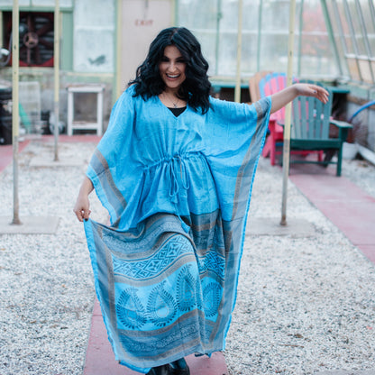A woman wearing an ice blue recycled Sari Long Kaftan.  She's paired it with a black thank top underneath and black boots. The Kaftan has peacock patterns on it.