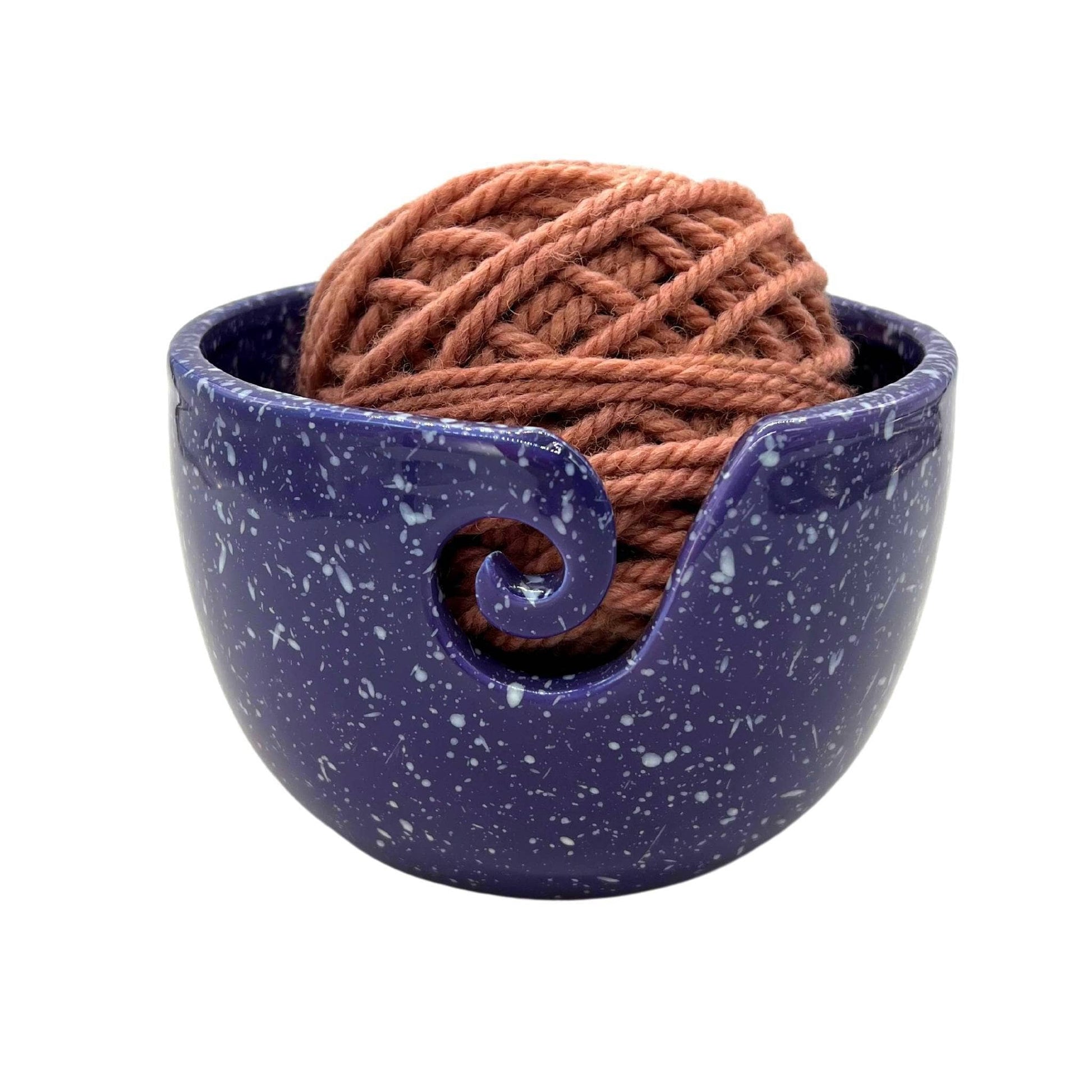 Purple with white speckled ceramic yarn bowl with salmon colored yarn inside of it on a white background