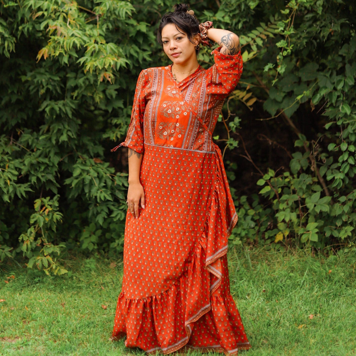 A model standing in a forest wearing a Burnt Red Floral Sari Wrap Dress. She has a matching Sari Silk Scrunchie on her wrist.