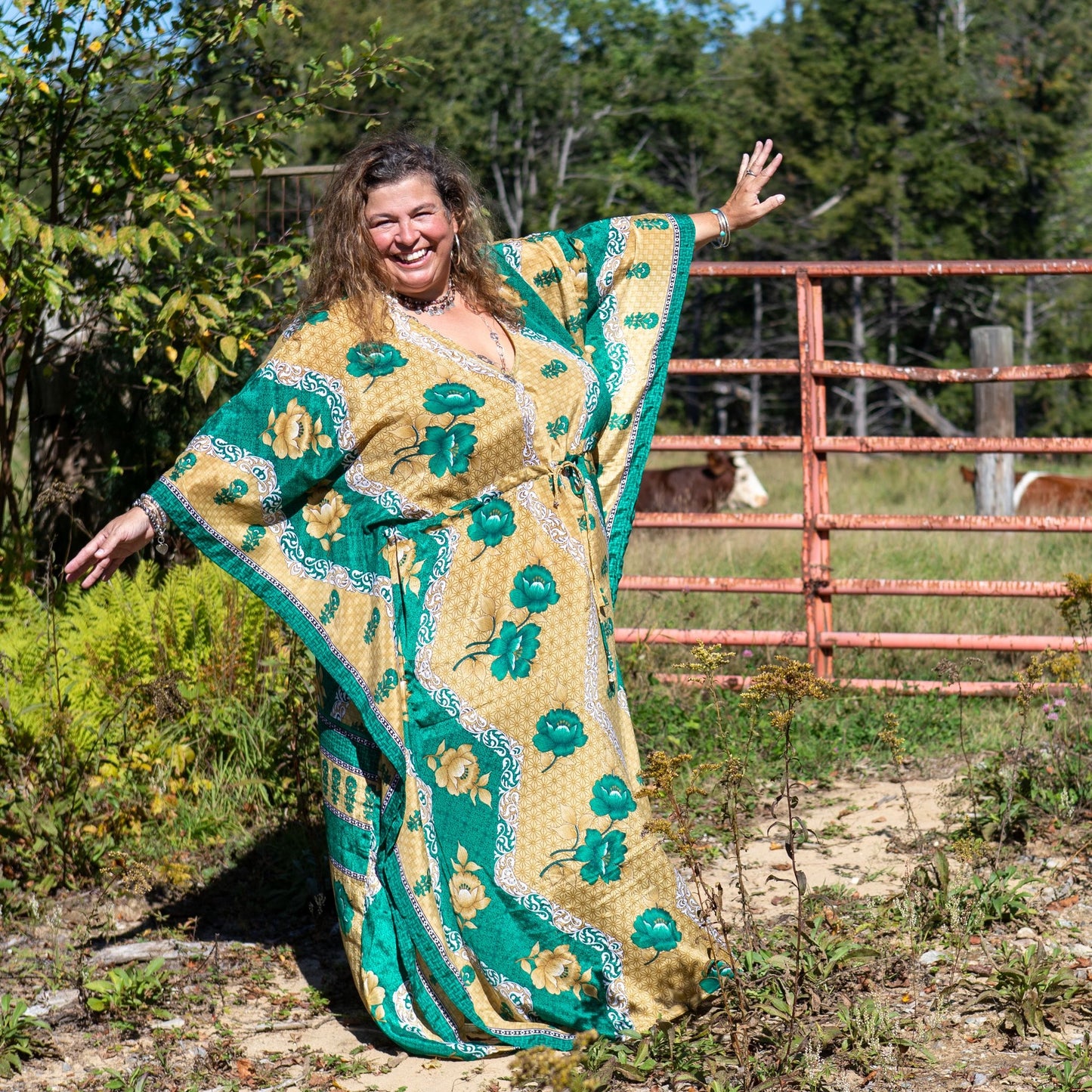 A Woman in a field wearing a Goddess size Aanya Adjustable Long Kaftan. This dress is made from reclaimed sari material and his primarily warm yellows and emerald greens. There are floral patterns on the edges.