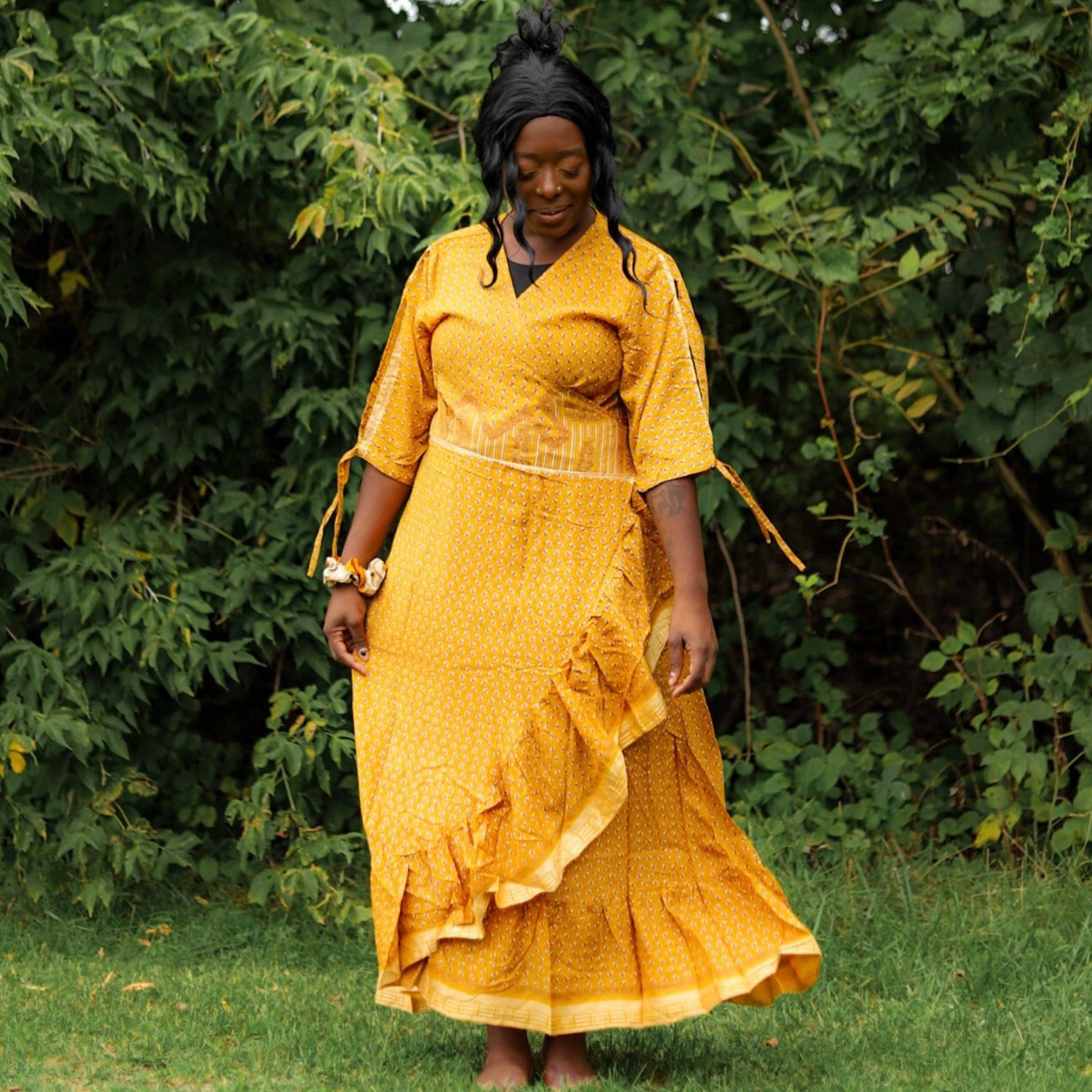 A Model standing in a forest wearing a Bright Yellow Zaria Wrap Dress. The Model is holding out the sides to show how flowy the fabric is.