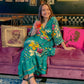 A model sitting in a bar holding a cocktail. She's wearing a Green Zaria Wrap Dress with Yellow, White and Pink flowers on it. She's sitting on a pink velvet couch.