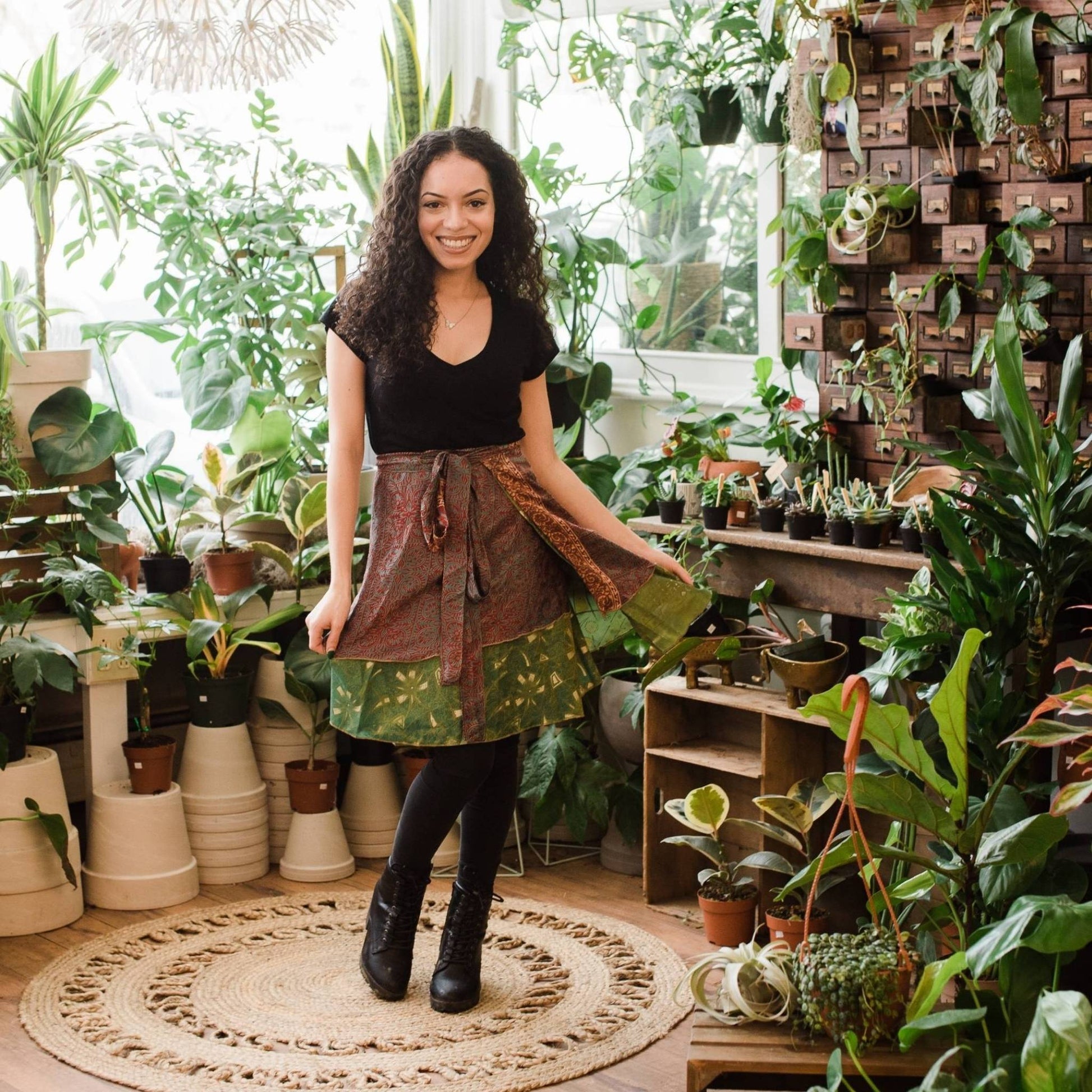 Petite model wearing a one of a kind mini sari wrap skirt in brown and green while standing in front of assorted potted plants.