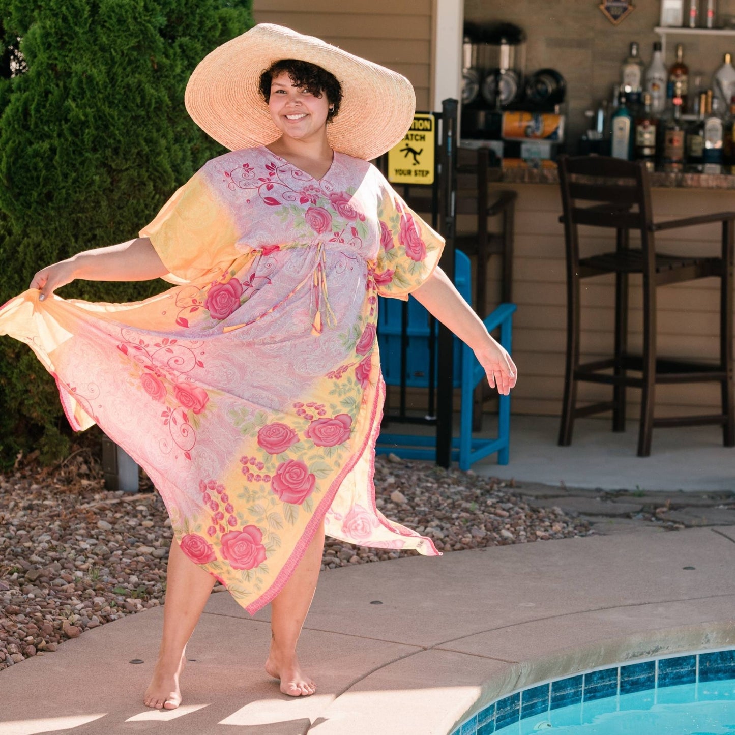 A woman by the pool wearing a light pink and yellow aanya adjustable kaftan. This dress has beautiful pink roses on it. She's paired the dress with a big sun hat.