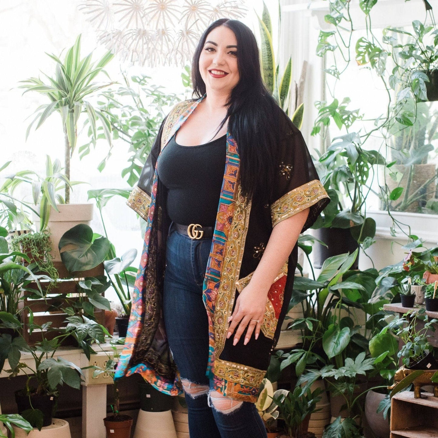 Plus size model is wearing a black sophia silk duster with a gold trim with potted plants in the background.