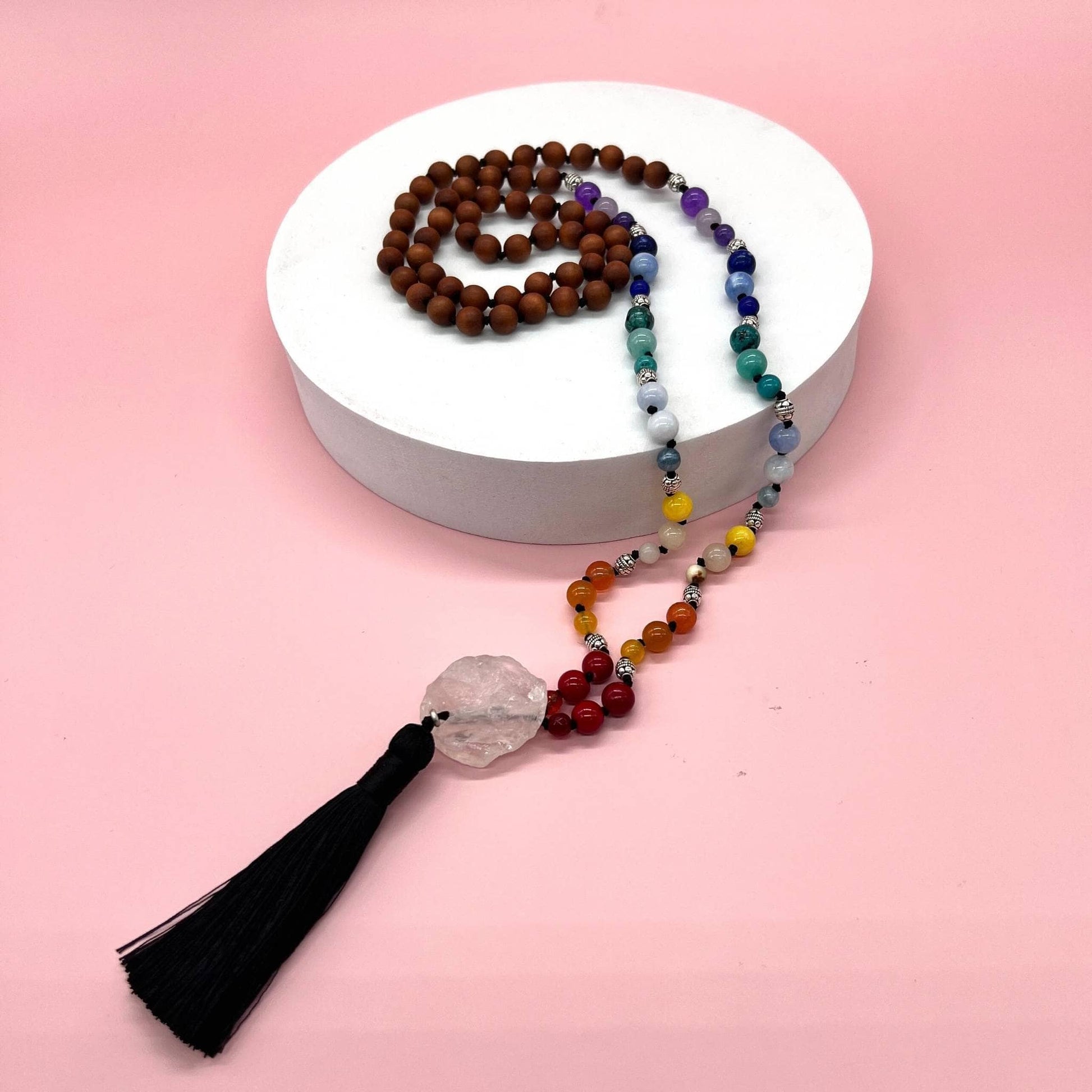 Chakra & Sandalwood Beaded Necklace is laying on a white flat cylinder on a pink backdrop.