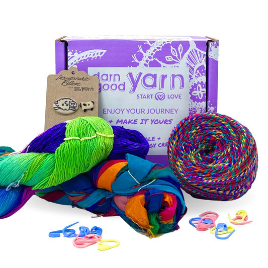 A Craft Sample Box on a white background. This box contains a sample of lace weight, Ribbon and Darn Good Sport Weight Twist Yarn. There are stitchable sheep buttons and pastel stitch markers in front of the box.