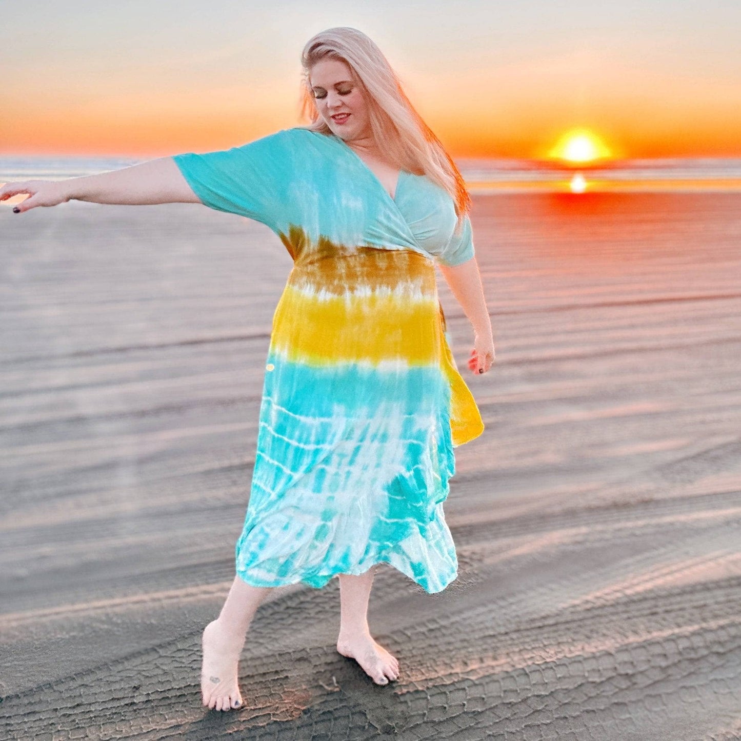 A blonde woman on a beach during sunset wearing a tie dye wrap dress. She's letting her toes run through the sand.