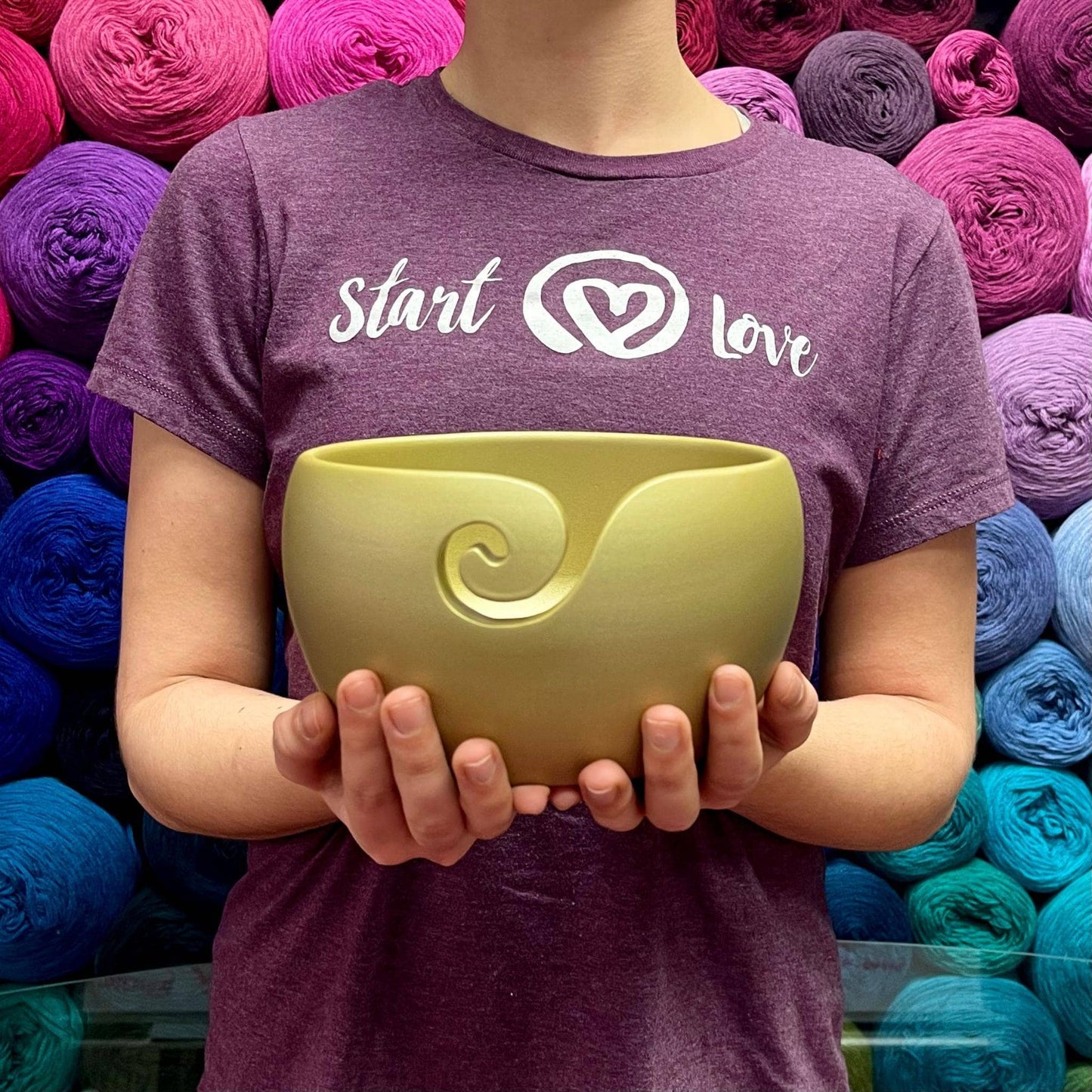 Model holding gold finished wooden yarn bowl while wearing purple start with love t-shirt and multicolor yarn background.