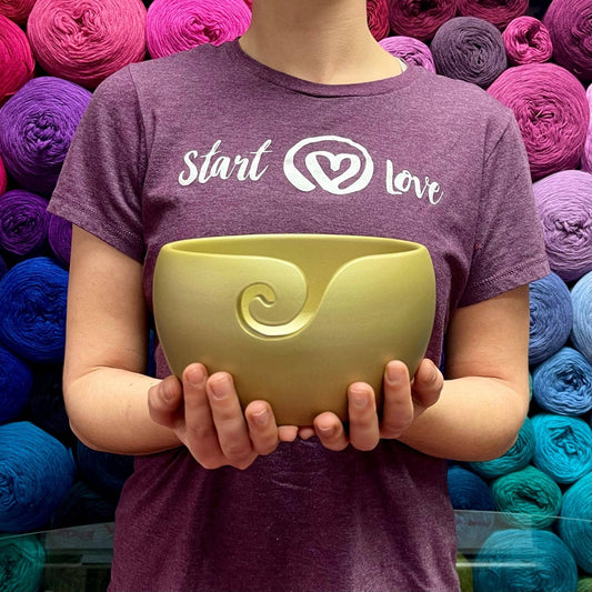 Model holding gold finished wooden yarn bowl while wearing purple start with love t-shirt and multicolor yarn background.