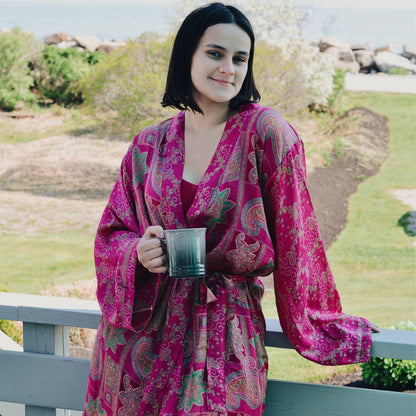 Model is standing outside on a deck holding coffee wearing a pink charlotte chic robe. 