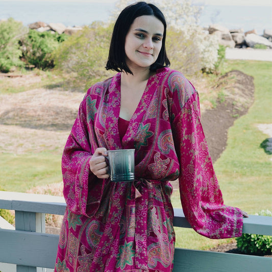 Model is standing outside on a deck holding coffee wearing a pink charlotte chic robe.