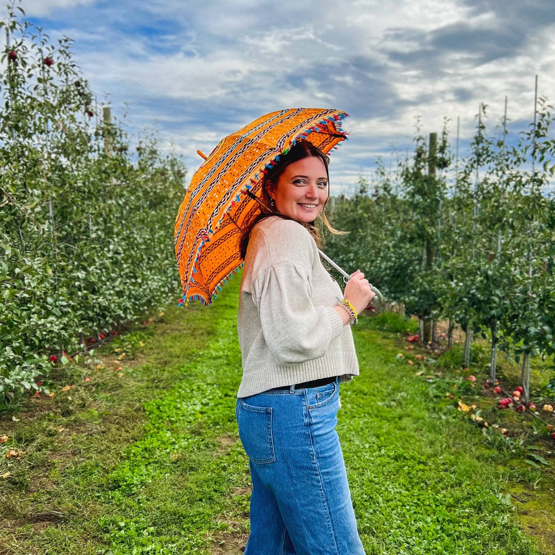 Model is standing in an apple orchard holding an orange Embellished Parasol Umbrella