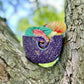 Ceramic yarn bowl purple with speckles filled with multiple skeins of yarn tucked into the nook of a tree with greenery in the background.