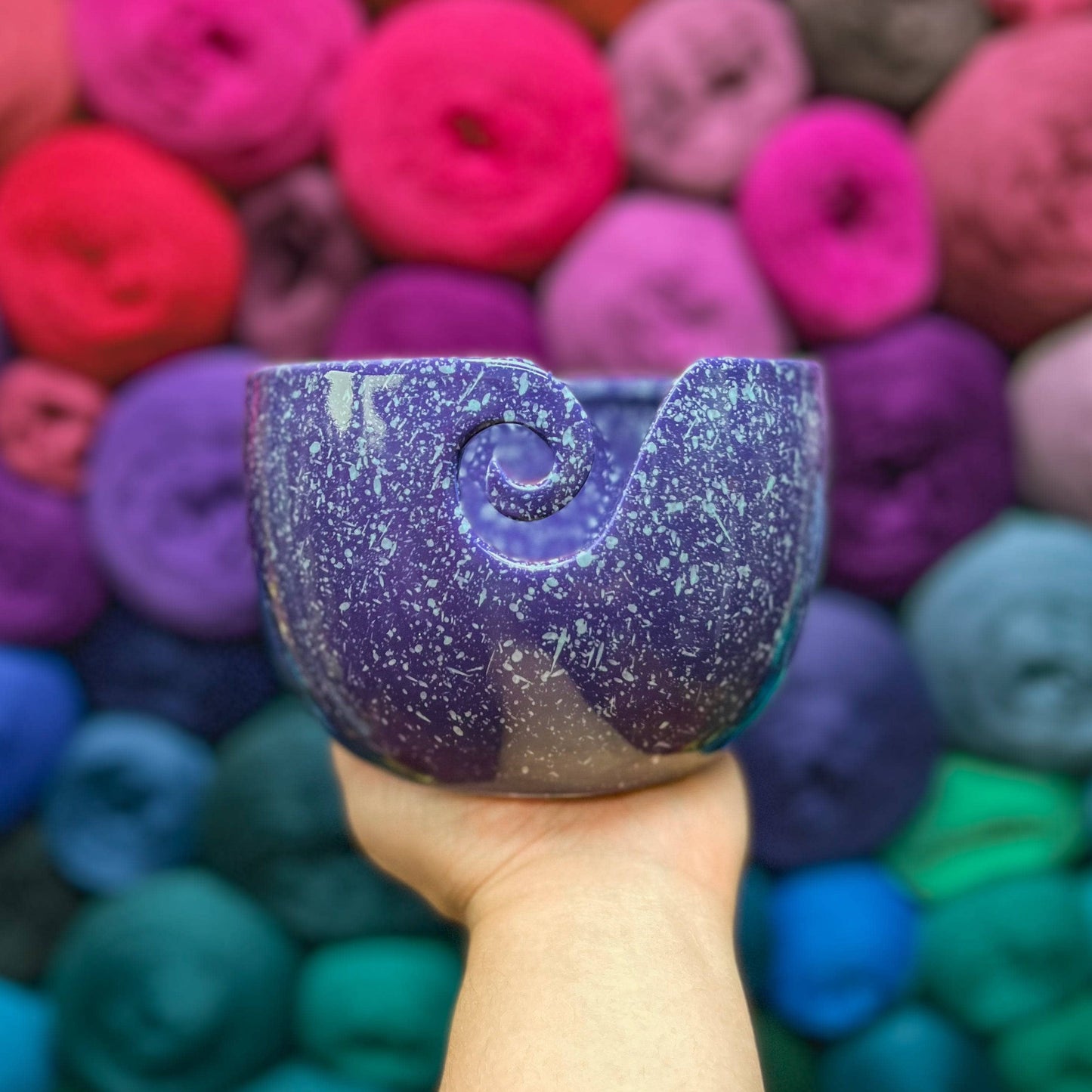 Hand holding up a purple ceramic yarn bowl with white speckles with multicolored yarn in the background.