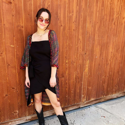 A woman standing outside in front of a wooden door. She's wearing a black dress and a maroon patterned Amara Short Duster