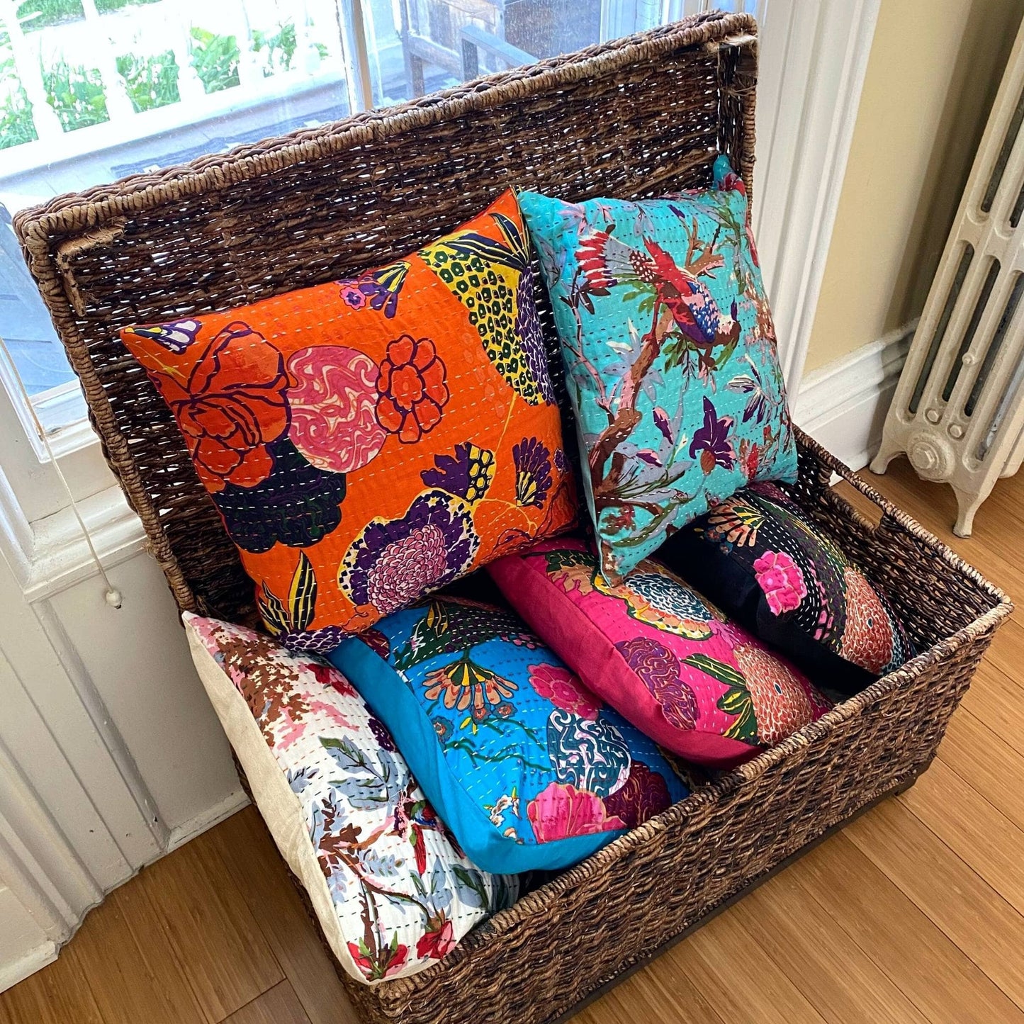 A stack of kantha pillow colors in a rectangular basket sitting in front of a window on a wooden floor.