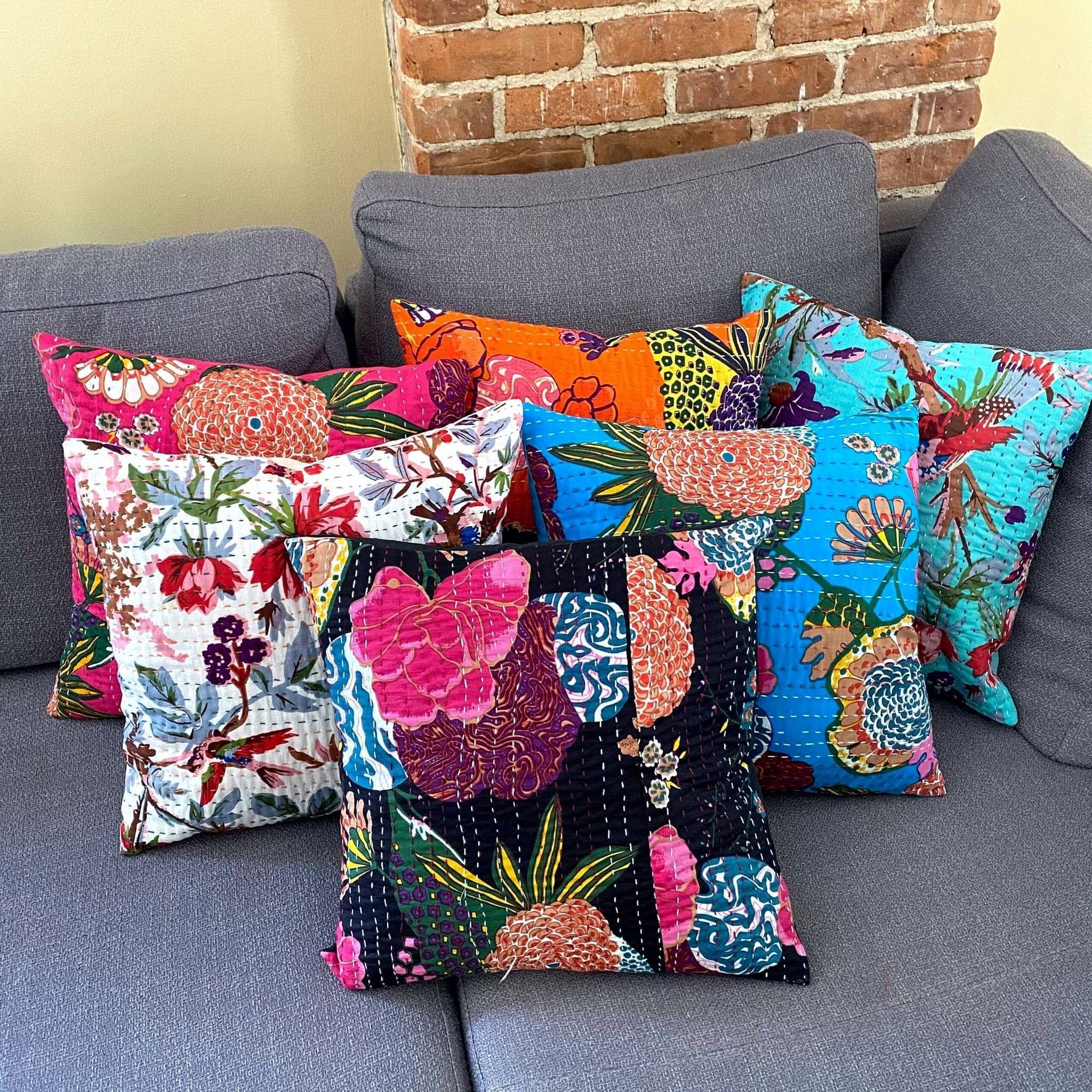 A pile of different Kantha pillow covers on a grey couch with a brick wall behind it.