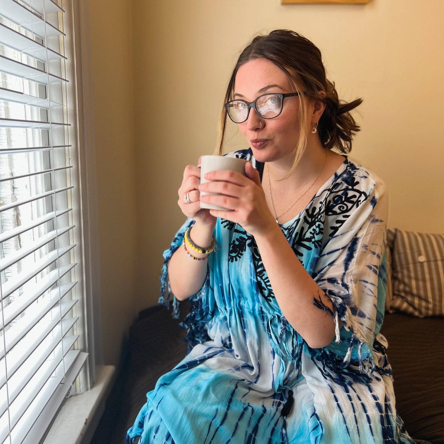 Woman in blue tie dye tunic about to take a sip of coffee in bed.