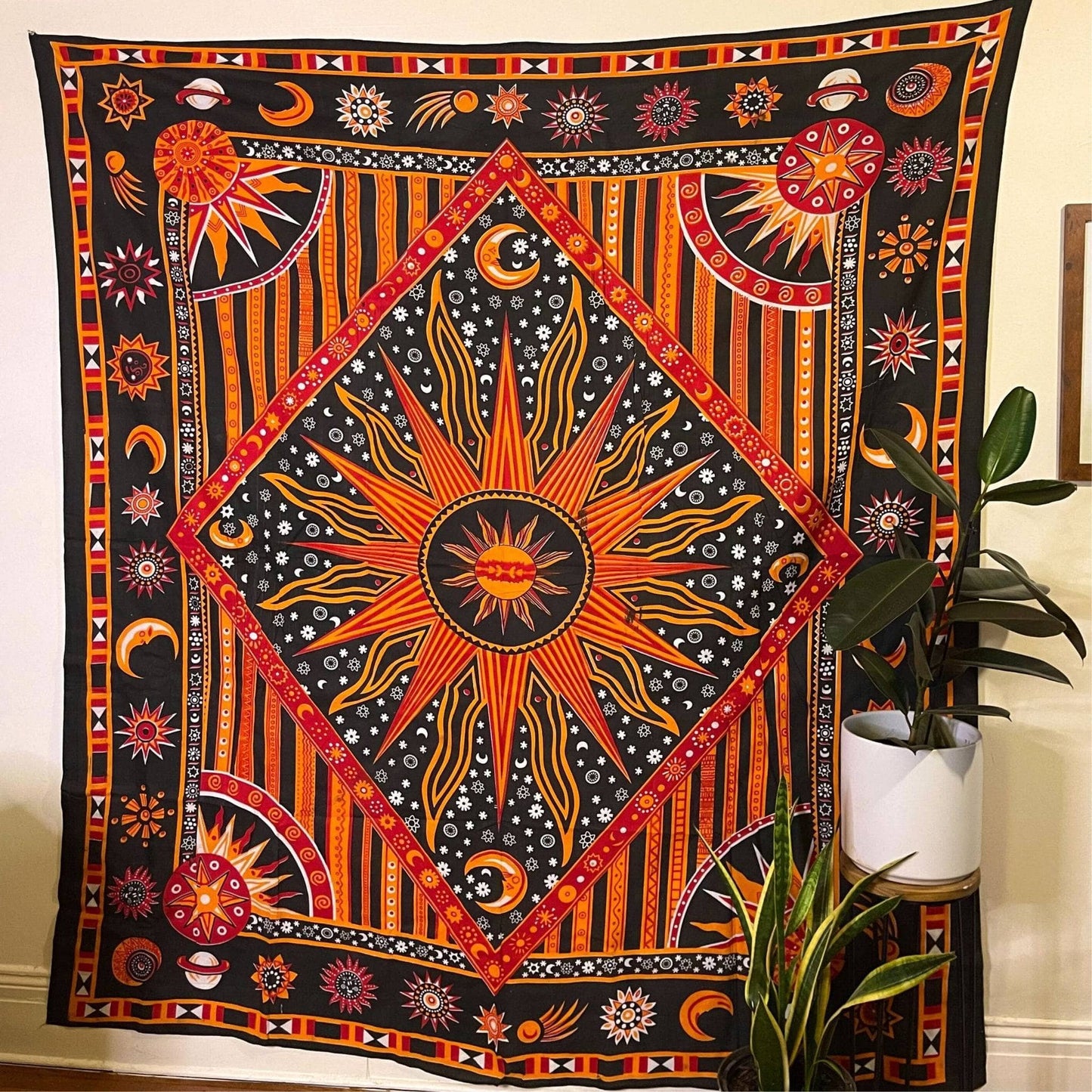 The Tapestry Psychedelic Celestial Sun hanging on a cream colored wall. There are plants sitting on the ground in front of it. This tapestry is a black background with bright oranges and reds mixed in, making a sun pattern in the center. There are stars, moons and suns around the edges.
