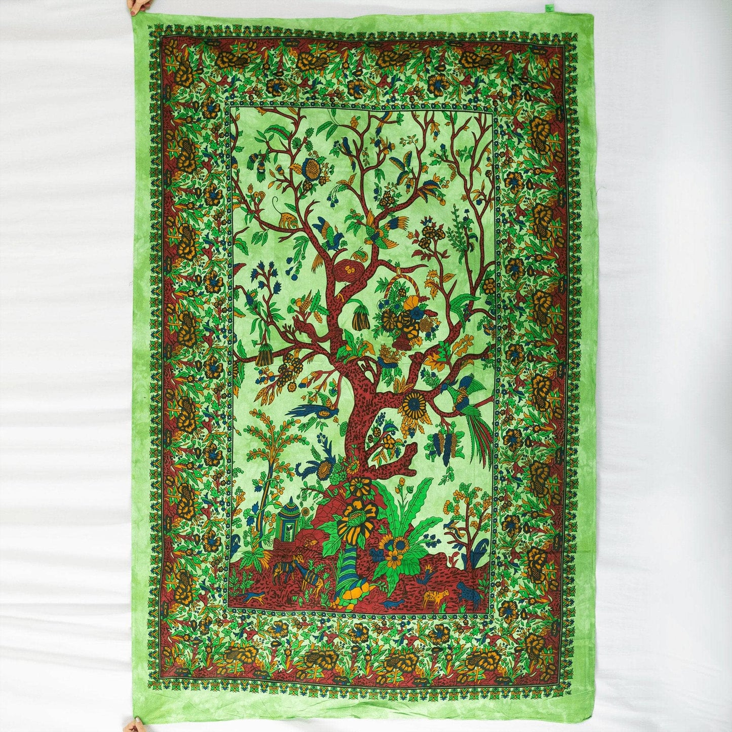 Tranquil tree tapestry hanging in front of a white background. Mainly green and brown with graphic of tree, birds, animals, and flowers.