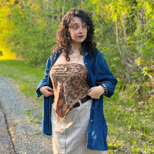 A girl standing on a gravel road wearing a sari silk bandana top. She's wearing a denim jacket over top and a button down skirt.. She's paired the look with boho jewelry