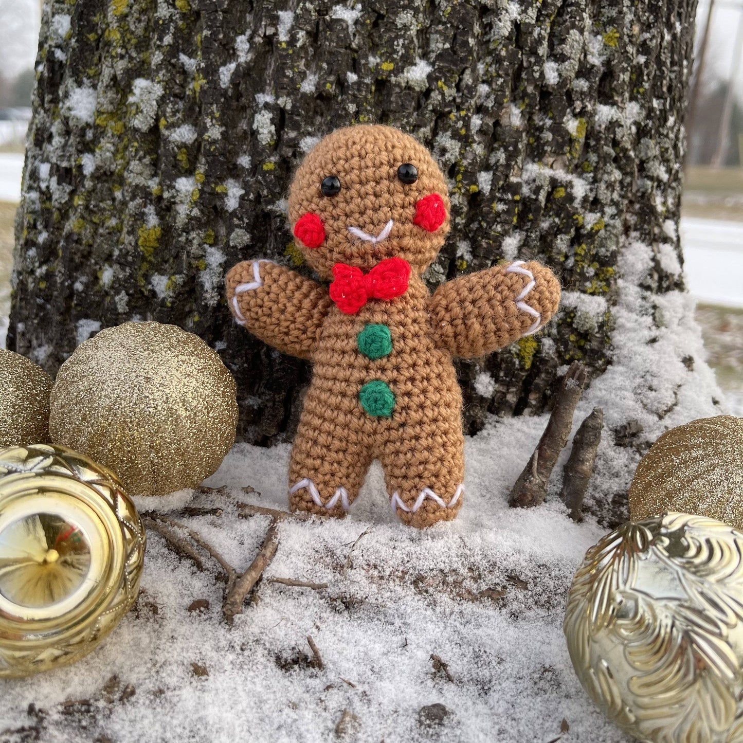 lifestyle shot of completed amigurumi gingerbread man standing in front of a snowy tree with christmas tree ball decorations around.